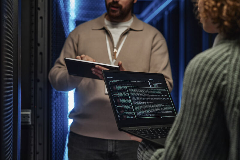 Two people in a data centre, viewing code on a laptop beside server racks with blue lighting.